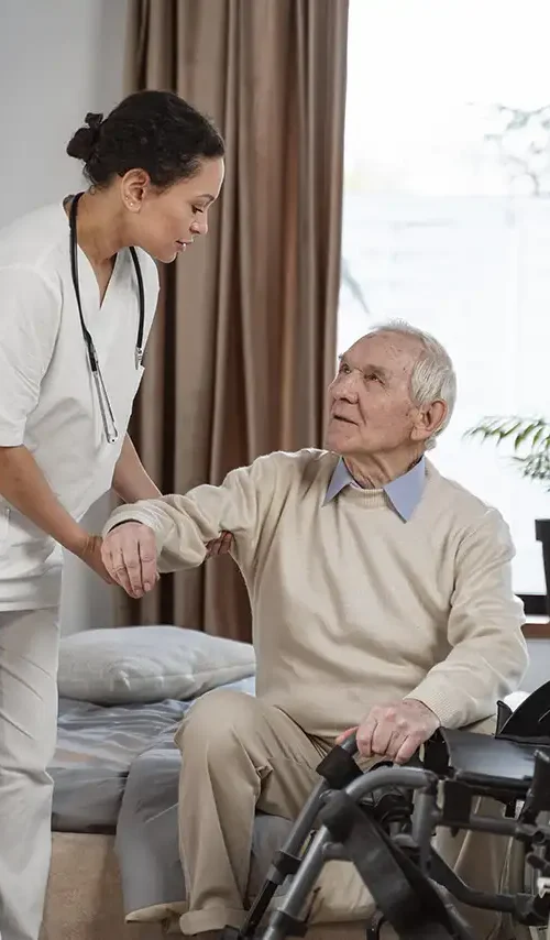 Nurse helping elderly man get into his wheelchair in his bedroom.
