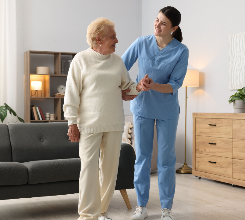A nurse assists an elderly woman in a cozy living room, providing care and support in a warm environment.