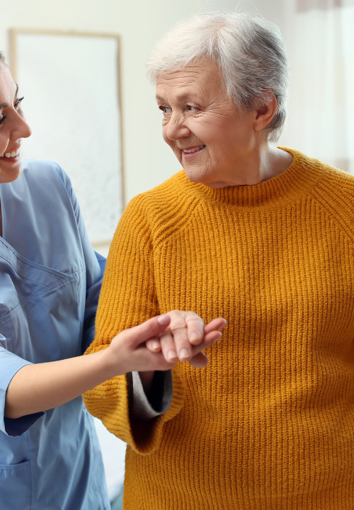 A caregiver gently supports a smiling senior woman as they walk together at home.