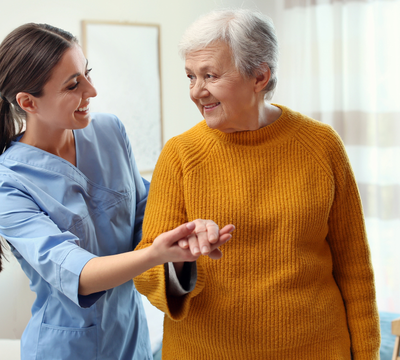 A caregiver gently supports a smiling senior woman as they walk together at home.