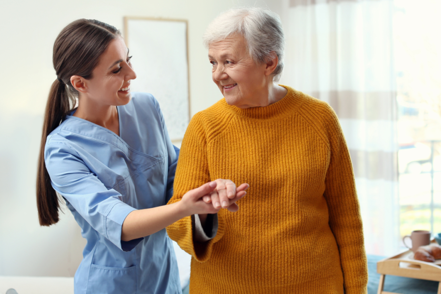 A caregiver gently supports a smiling senior woman as they walk together at home.